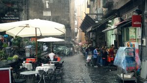 an image of a city in Italy in heavy rainfall, there are large umbrellas over tables and chairs, tarpaulins over street furniture, and a small crowd of people gathered in a shop entrance
