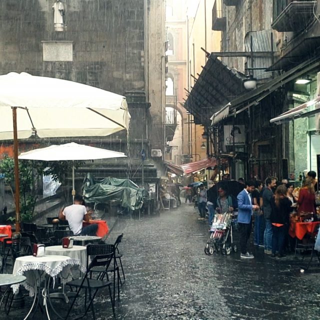 an image of a city in Italy in heavy rainfall, there are large umbrellas over tables and chairs, tarpaulins over street furniture, and a small crowd of people gathered in a shop entrance