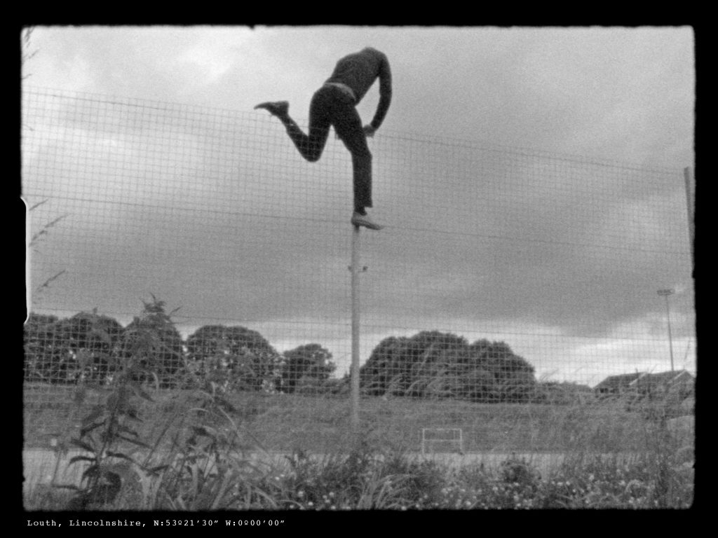 A black and white film still, showing a man climbing over a thin fence, approximately 3 metres tall. He has reached the top and has one leg over each side.