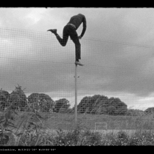 A black and white film still, showing a man climbing over a thin fence, approximately 3 metres tall. He has reached the top and has one leg over each side.