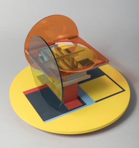 An abstract sculpture on a light grey background. It includes a large pastel yellow circle base, painted with navy blue squares. The sculpture is an assemblage of clear coloured perspex sheets and small pieces of MDF, in circle and square shapes. The colours are mainly orange, blue and yellow.