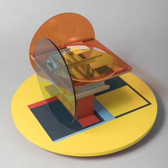 An abstract sculpture on a light grey background. It includes a large pastel yellow circle base, painted with navy blue squares. The sculpture is an assemblage of clear coloured perspex sheets and small pieces of MDF, in circle and square shapes. The colours are mainly orange, blue and yellow.