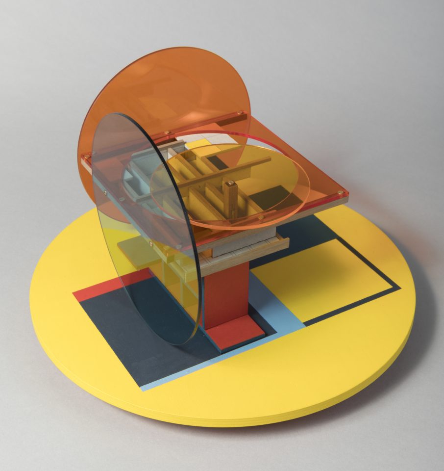 An abstract sculpture on a light grey background. It includes a large pastel yellow circle base, painted with navy blue squares. The sculpture is an assemblage of clear coloured perspex sheets and small pieces of MDF, in circle and square shapes. The colours are mainly orange, blue and yellow.