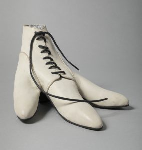 a large white leather handmade shoe with black laces sits on a grey background. Made for a bird rather than a human, it splits into three large toes.