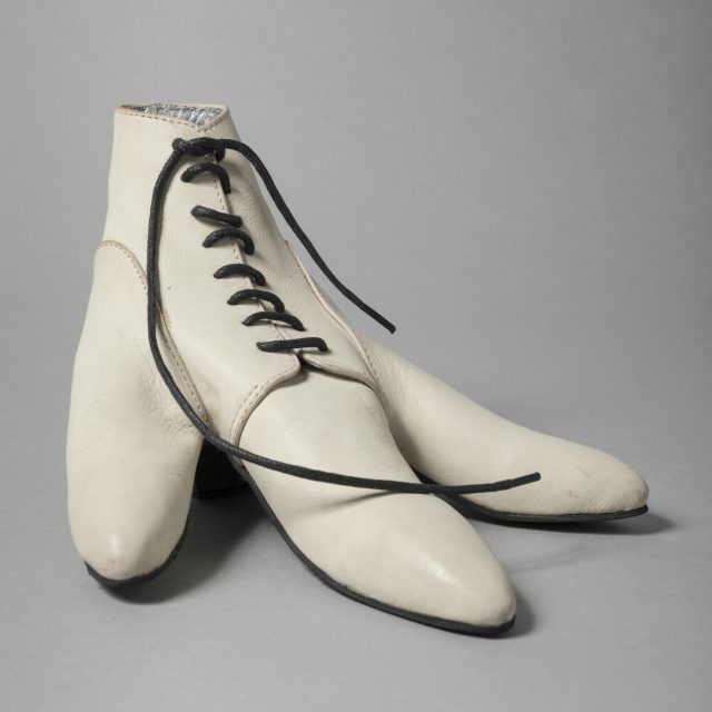 a large white leather handmade shoe with black laces sits on a grey background. Made for a bird rather than a human, it splits into three large toes.