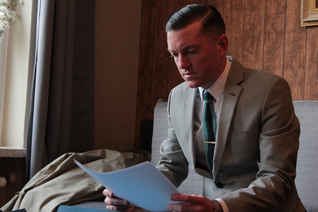A film still, in colour, showing a white man with short, slicked back cropped hair, reading a document. He is wearing a light brown suit and blue tie.