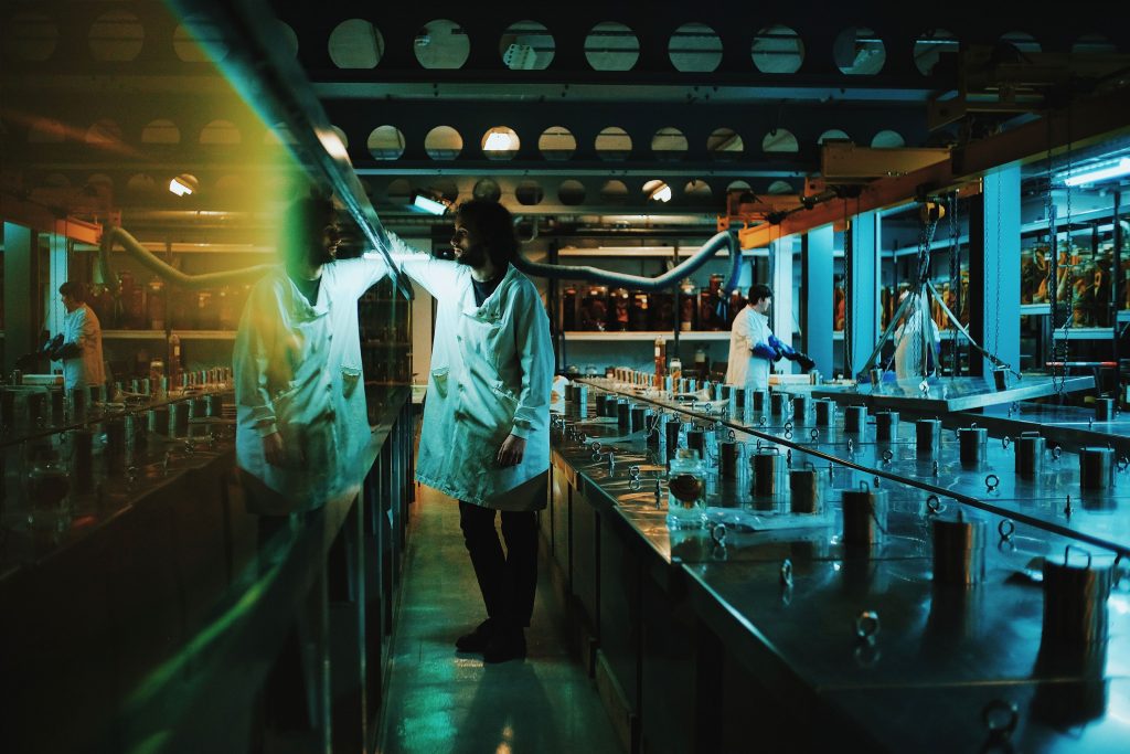 A still from a video, showing a person with a beard and shoulder length hair, in a white labcoat. They are looking into a glass chamber, which is obscured by reflection.