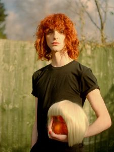A picture of a person stood in front of a fence. They have shoulder length red curly hair cut with a fringe. In their left hand they are holding a blonde wig