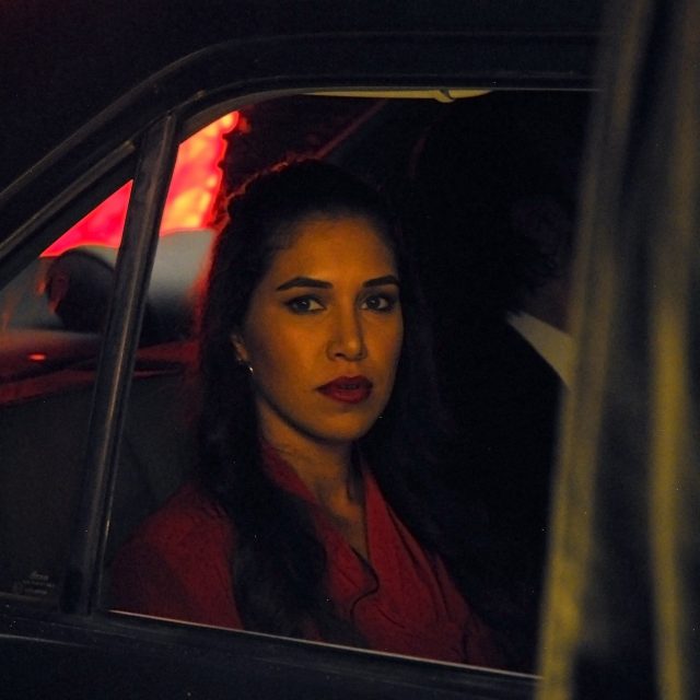 A film still, showing a woman looking out of a car window. From the lighting we can assume it is nightime