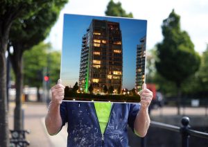 a photograph of a person, wearing a blue hoody with paint on it. They are holding up an image of a tower block, which obscures their face.