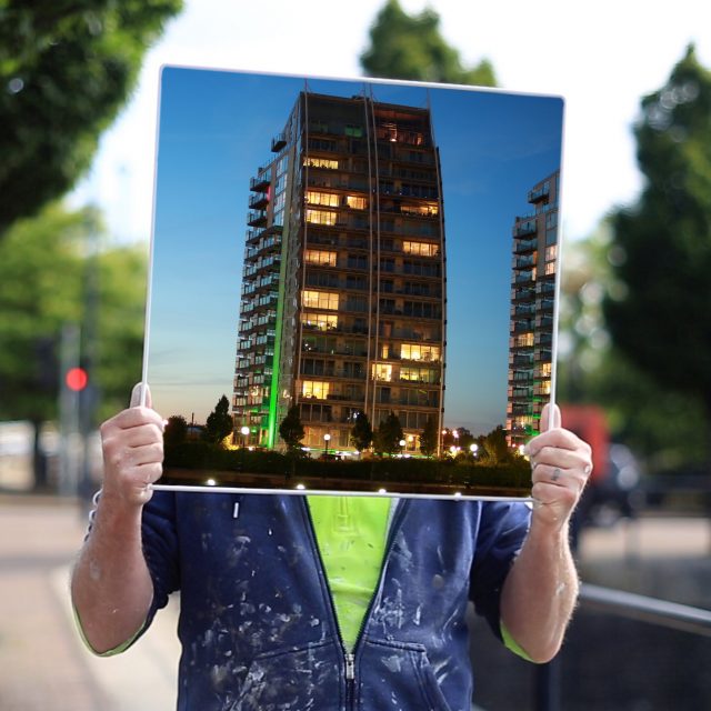 a photograph of a person, wearing a blue hoody with paint on it. They are holding up an image of a tower block, which obscures their face.