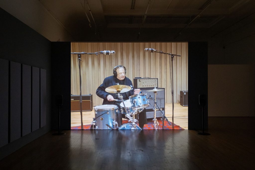 A photograph of a projection in a dark room in Salford Museum and Art Gallery. The footage shows an older white man playing the drums inside a professional music recording studio.