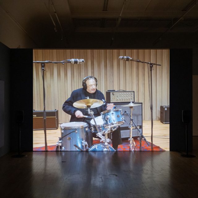 A photograph of a projection in a dark room in Salford Museum and Art Gallery. The footage shows an older white man playing the drums inside a professional music recording studio.