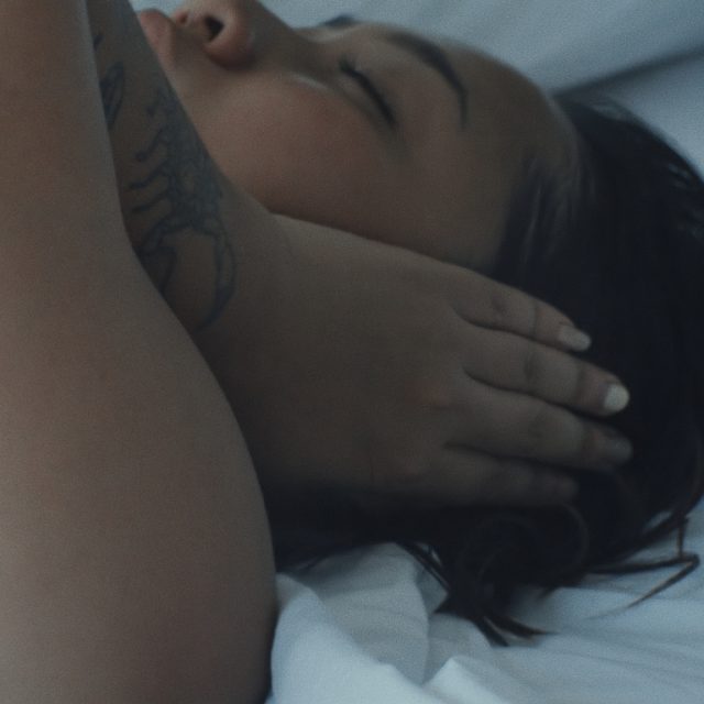 a still from a video, showing a close-up of a person lying on a bed. They are covering their left ear with their left hand