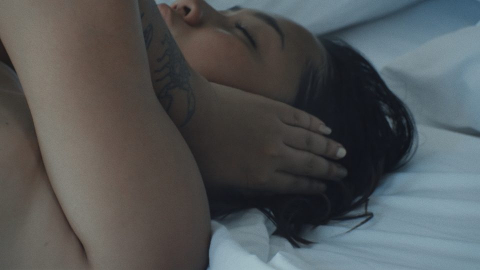 a still from a video, showing a close-up of a person lying on a bed. They are covering their left ear with their left hand