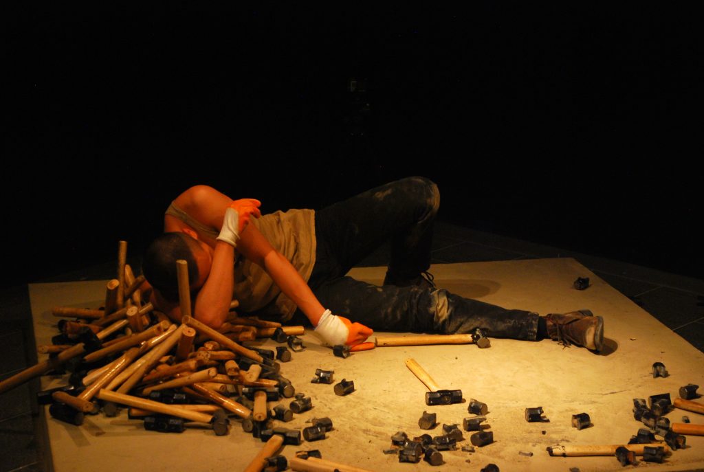 a man wearing orange work gloves lies exhausted on a concrete plinth. He is surrounded by lots of hammers whose heads have been smashed to pieces. In his hand he hold a single unbroken hammer.