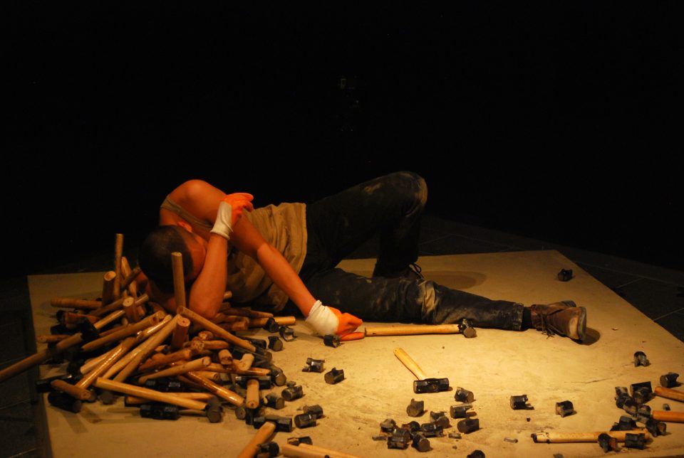 a man wearing orange work gloves lies exhausted on a concrete plinth. He is surrounded by lots of hammers whose heads have been smashed to pieces. In his hand he hold a single unbroken hammer.