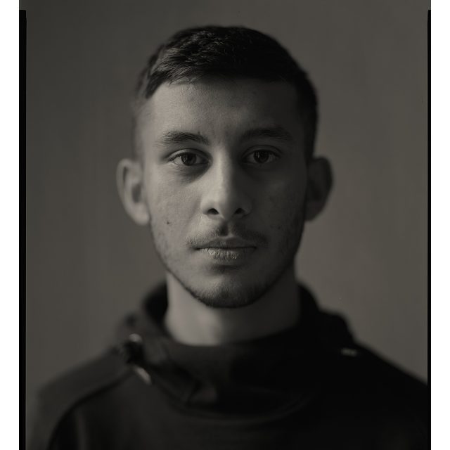 A black and white photograph of a younger person. They have short dark hair and a short beard and moustache. They're looking straight towards the camera.