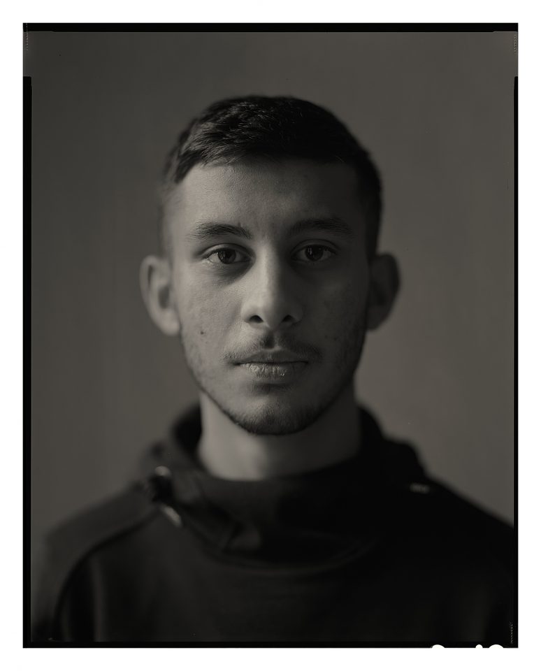 A black and white photograph of a younger person. They have short dark hair and a short beard and moustache. They're looking straight towards the camera.