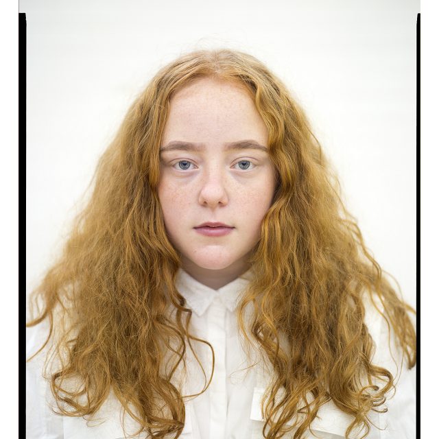 an image of a young person, with long way auburn hair. She looks towards the camera.
