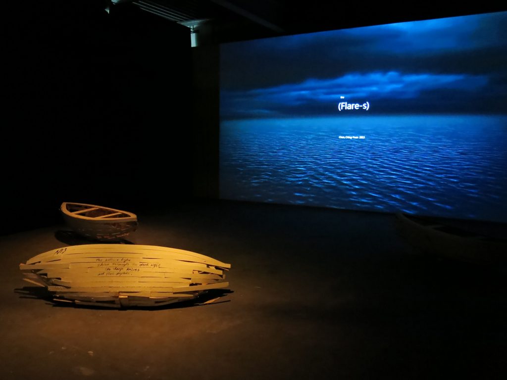 A paper boat and a real boat in front of a blue projection.