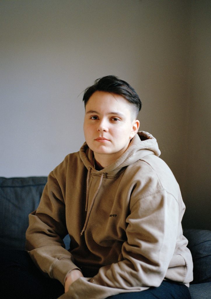 A colour photography showing a white person from the waist up. They dark hair which is shaved at the sides and they're sat on a black sofa wearing a beige, oversized hoodie.