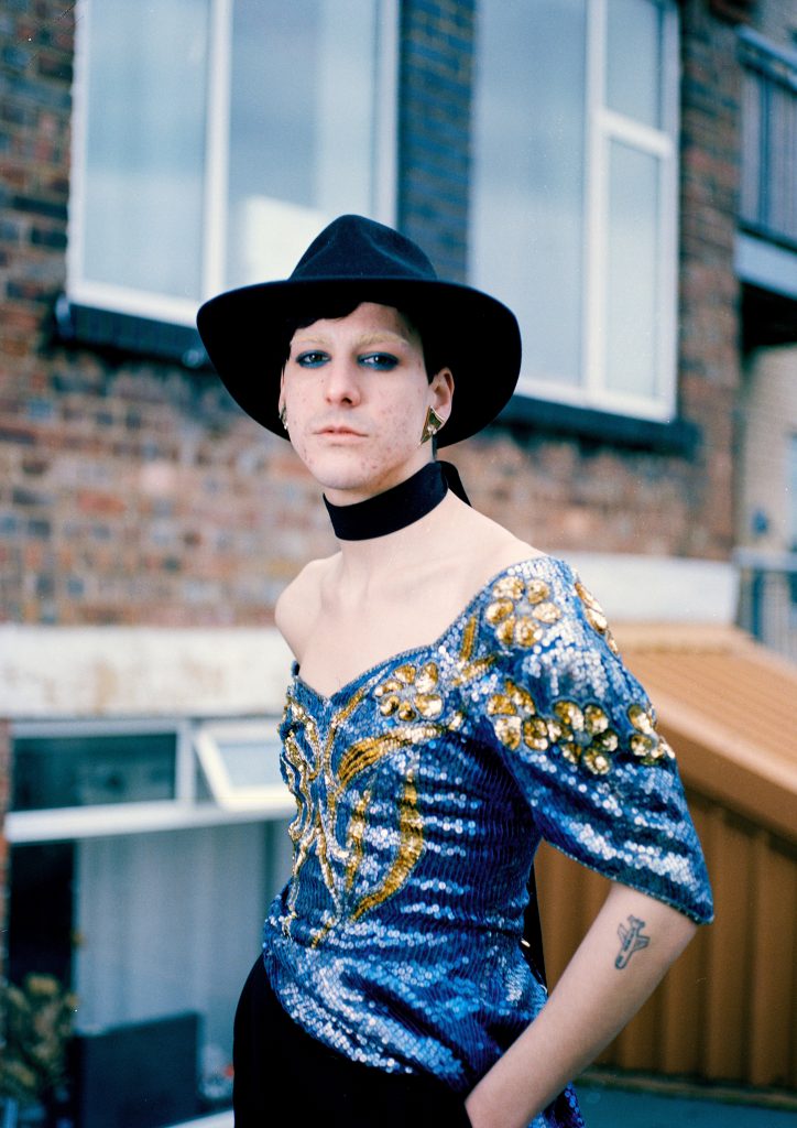 A colour photography shows a white person from the waist up, stood outside in front of a brick built house. They are wearing a sequin, off the shoulder blue and gold top, a wide black choaker, and a black hat. They are wearing blye eyeshadow, and large geometric gold earings.