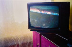 A CRT TV sites on the edge of a black and pink cabinet. Behind, a thin light net curtain allows a soft glow of light into the room. The TV screen is paused, the bottom half is black, in the upper half a pair of eyes and hat are just visible.