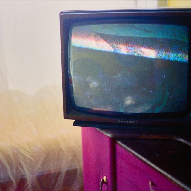 A CRT TV sites on the edge of a black and pink cabinet. Behind, a thin light net curtain allows a soft glow of light into the room. The TV screen is paused, the bottom half is black, in the upper half a pair of eyes and hat are just visible.