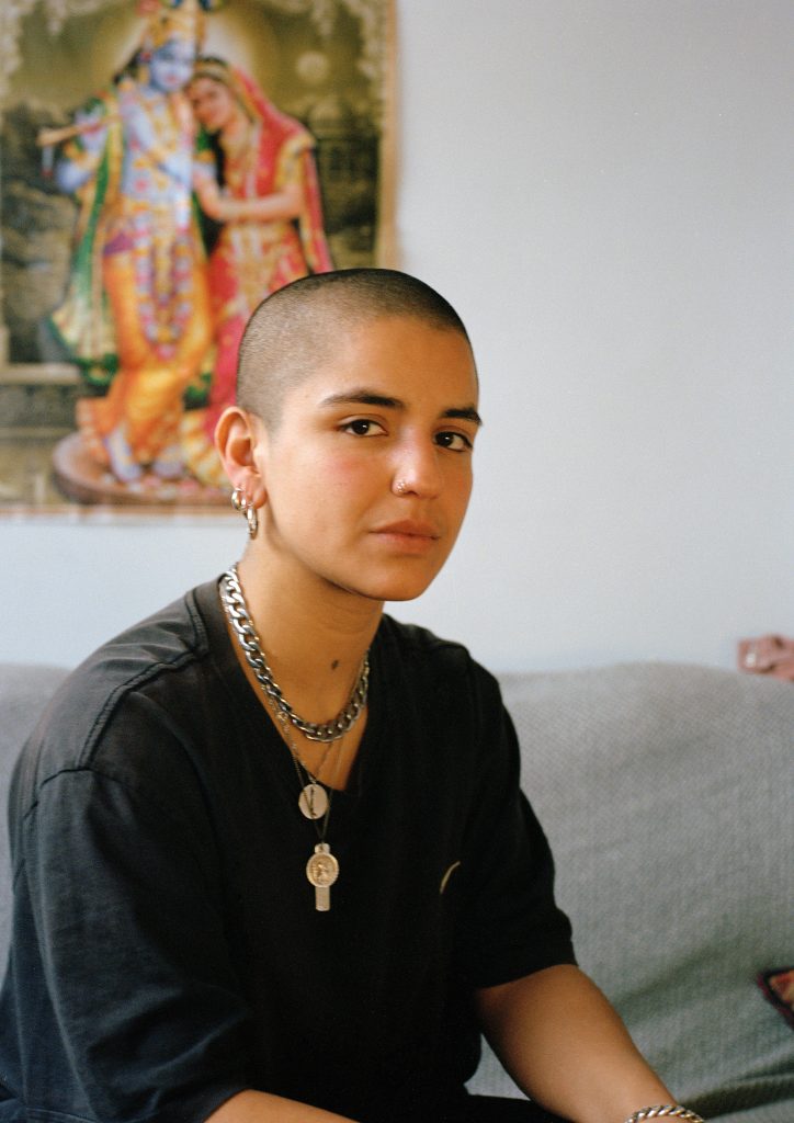 A colour photograph shows a person from the waist up, sat inside on a grey sofa. They have a shaved head and wear layered necklaces over a black tshirt. Behind them on the wall is a poster depicting Radha-Krishna.
