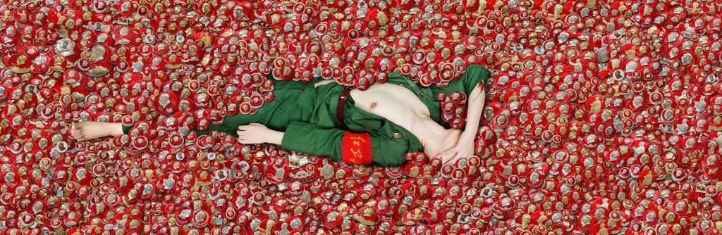 A woman in a green soldier's uniform lies with her breasts exposed in a pile of red medallions.