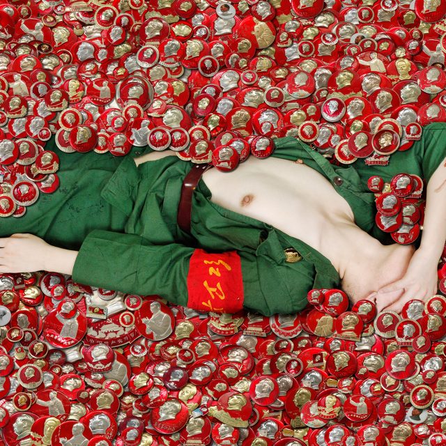A woman in a green soldier's uniform lies with her breasts exposed in a pile of red medallions.