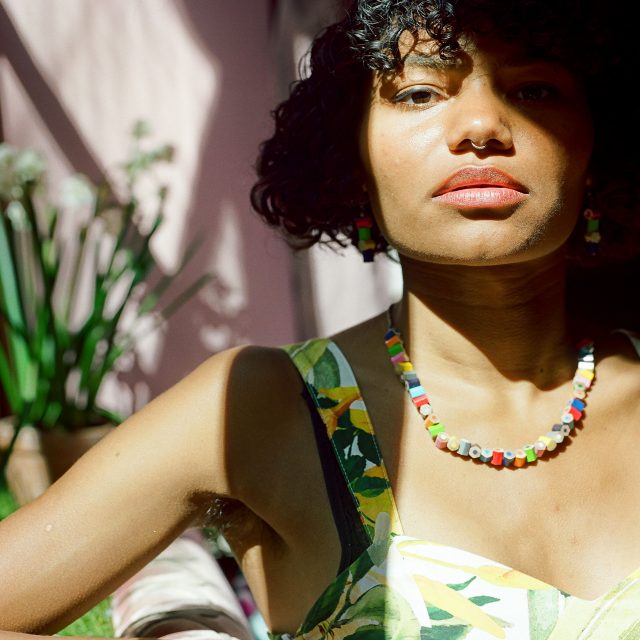 A colour photograph shows a black person. They have curly hair styled in a bob and are wearing a necklace and earings made from coloured pencil segments, and a dress with a green and yellow floral pattern. They have their nose piereced and visible armpit hair. Their right eye is half obscured by shadow.