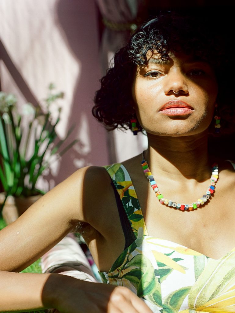 A colour photograph shows a black person from the chest up. They have curly hair styled in a bob and are wearing a necklace and earings made from coloured pencil segments, and a white dress with a green and yellow floral pattern. They have their nose piereced and visible armpit hair. Their right eye is half obscured by shadow.