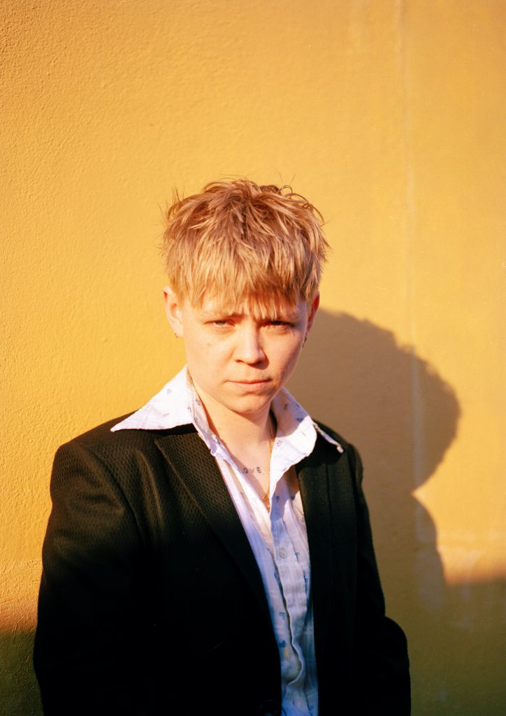 A bright colour photography shows a white person from the waist up, posed against a yellow backgroud. They have short blond hair, and they are wearing a button up shirt with a statement collar, under a black blazer jacket.