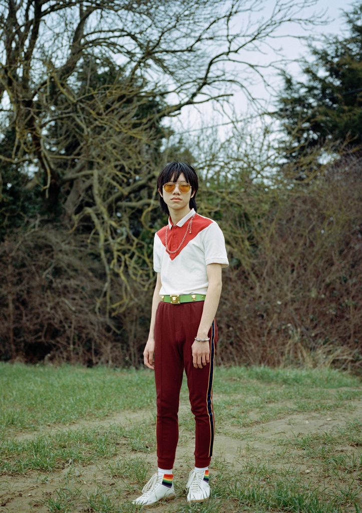 A full body colour photography showing an asian person in red trousers, a red and white graphic button-up shirt, and statement orange-tinted glasses stands in a field facing the camera.