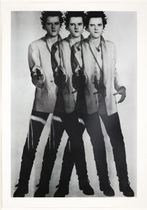 A black and white print shows the artist stood in overlaping triplicate, with his feet apart, holding a pistol at waist height, pointed towards the viewer. He is dressed as Sid Vicious, impersonating Elvis.