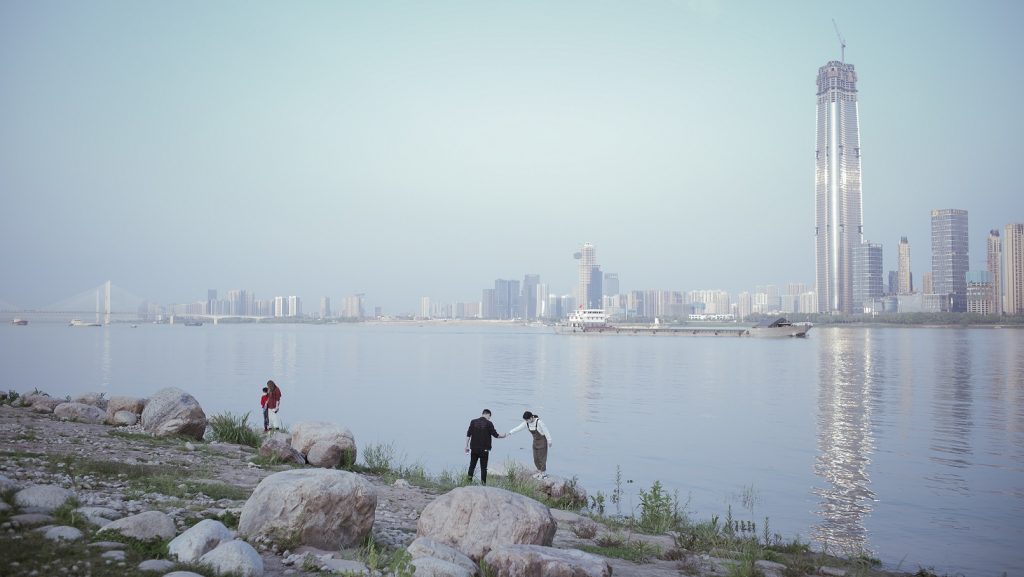 a light blue sky and sea, a rocky shore in the foreground. The wide image shows an urban skyline in Wuhan, China, with tall buildings and skyscrapers. In middle distance on the shore are a few small human  figures, two in the centre reach out and hold hands.
