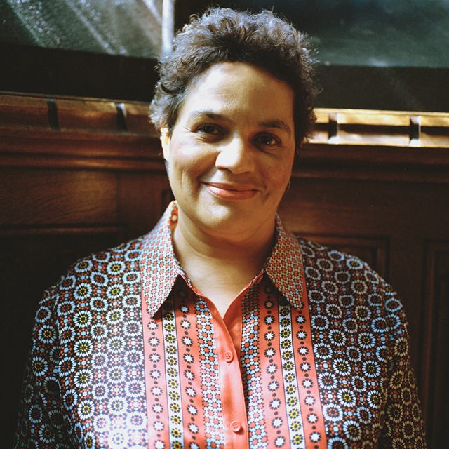 A digital photograph shows Jackie Kay, a black woman with short hair, wearing a black and red paterned shirt.