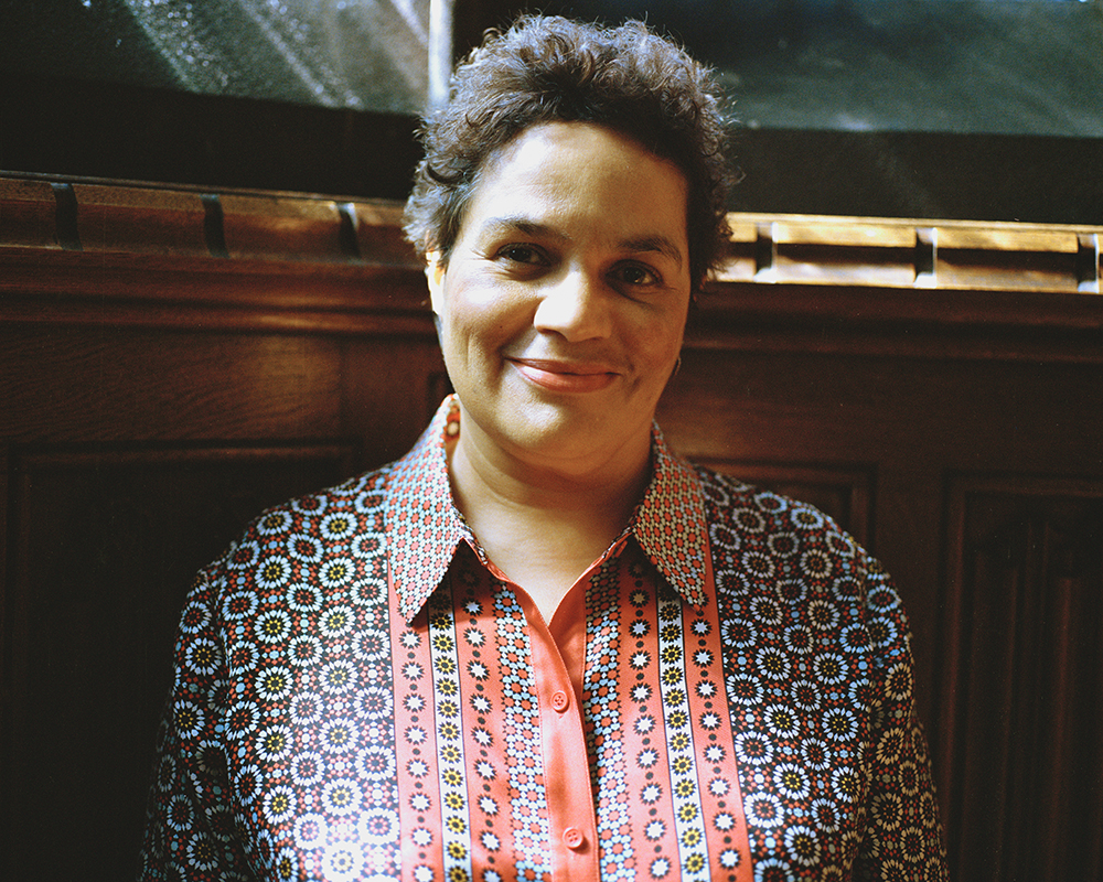 A digital photograph shows Jackie Kay, a black woman with short hair, wearing a black and red paterned shirt.