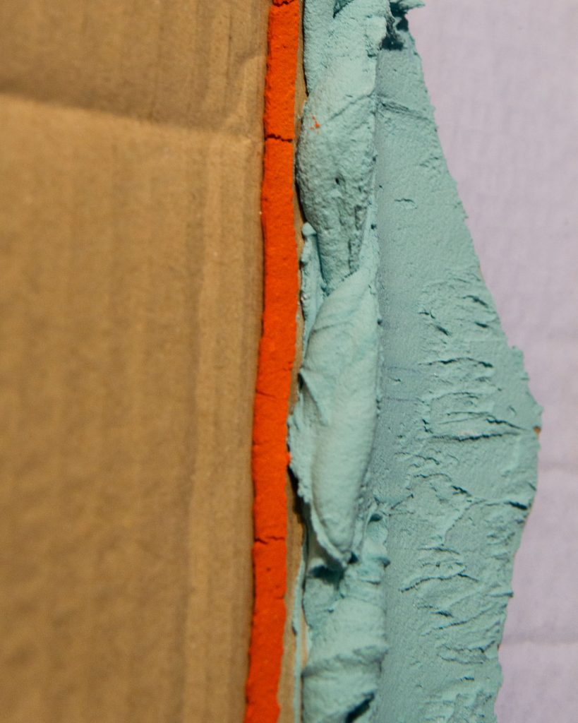 A close-up of the righthand object, course desaturated orange and cyan paint spills out from behind a cardboard curtain.