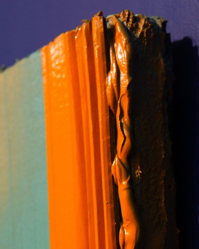 A close-up of the left-hand object. Orange paint is pressed between cardboard walls, it spills out, still layered like a globular stack of paper.