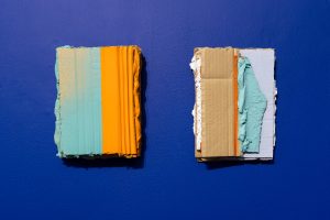 2 portrait orientated rectangles, in an almost 4 by 3 format. The left one has a subdued cyan tone on its left side, and a desaturated orange on the right. The righthand object is cyan and white on its right side, but bare cardboard on its left. Both are textured with thick paint and cardboard, hung on a blue wall.