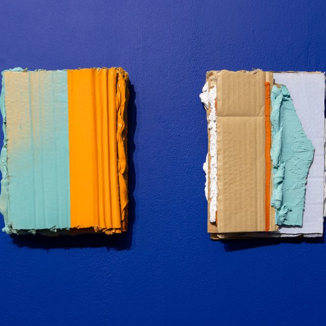 2 portrait orientated rectangles, in an almost 4 by 3 format. The left one has a subdued cyan tone on its left side, and a desaturated orange on the right. The righthand object is cyan and white on its right side, but bare cardboard on its left. Both are textured with thick paint and cardboard, hung on a blue wall.