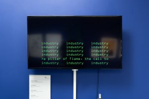 A large flat-screen television on a blue wall. The screen has green blocky text upon it, like you'd see in old movies where they hack computers - think the matrix. The screen reads: industry, a pillar of flame, the call to industry