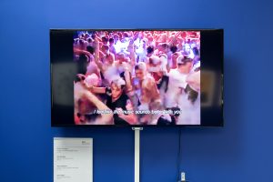 A large flat-screen television on a blue wall. The video on the screen depicts a club party, with everyone covered in bubbly foam. There are subtitles on the screen reading: "I feel like the music sounds better with you"