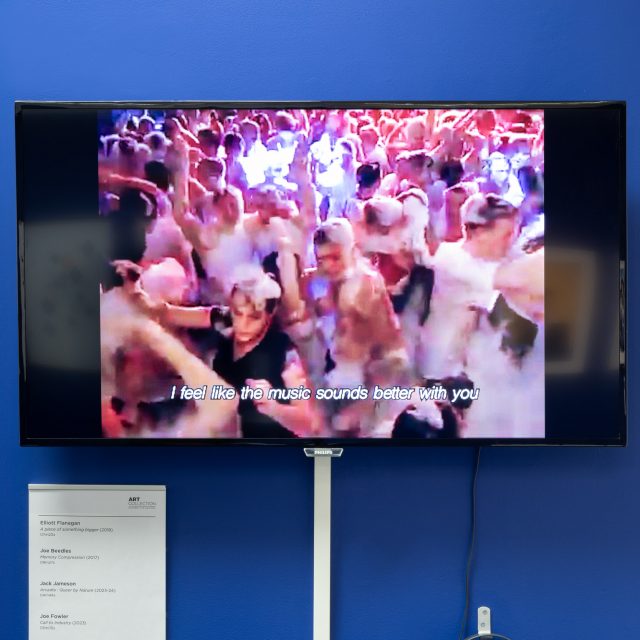 A large flat-screen television on a blue wall. The video on the screen depicts a club party, with everyone covered in bubbly foam. There are subtitles on the screen reading: "I feel like the music sounds better with you"