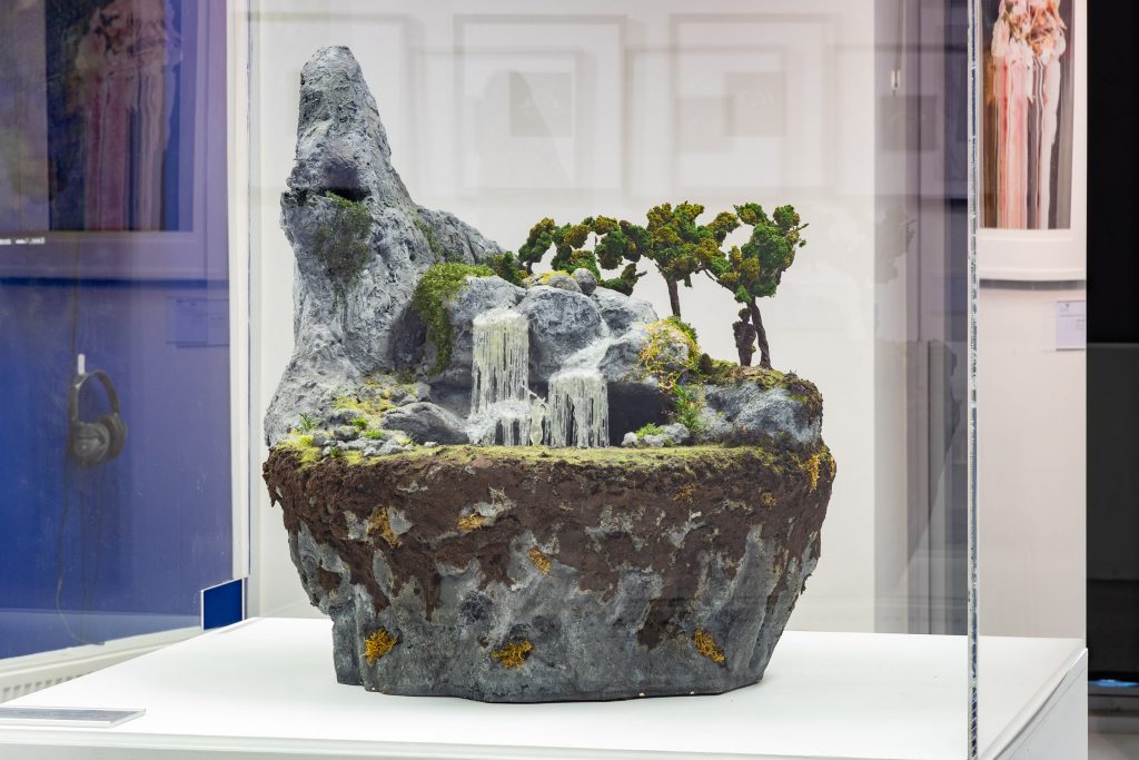 A large sculpture, a floating island. A lower rocky layer, followed by soil, then grass, then a whimsical scene. 3 figures - one perching on an outstretched cliff outside of a cave. The next half-submerged in the water below a waterfall running through the center of the sculpture. and the last amongst the trees toward the back side of this diorama.