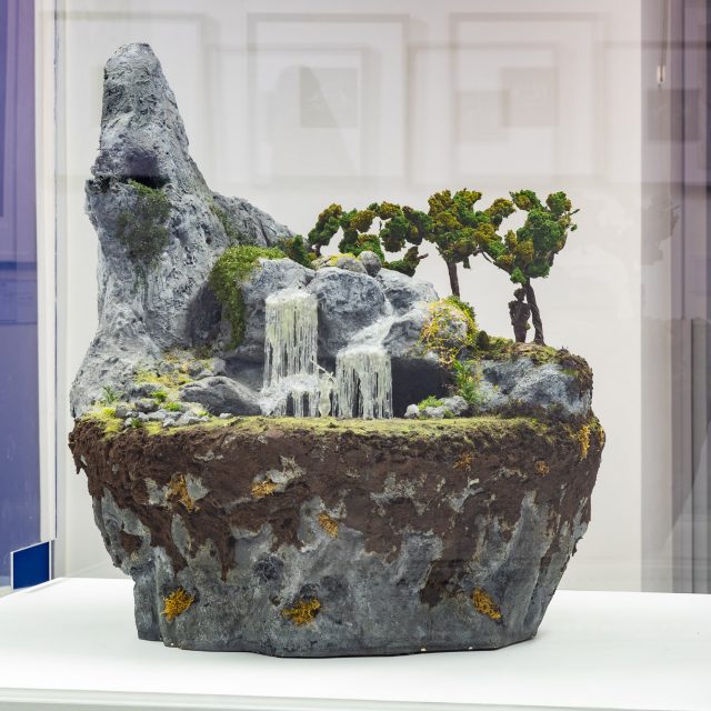 A large sculpture, a floating island. A lower rocky layer, followed by soil, then grass, then a whimsical scene. 3 figures - one perching on an outstretched cliff outside of a cave. The next half-submerged in the water below a waterfall running through the center of the sculpture. and the last amongst the trees toward the back side of this diorama.