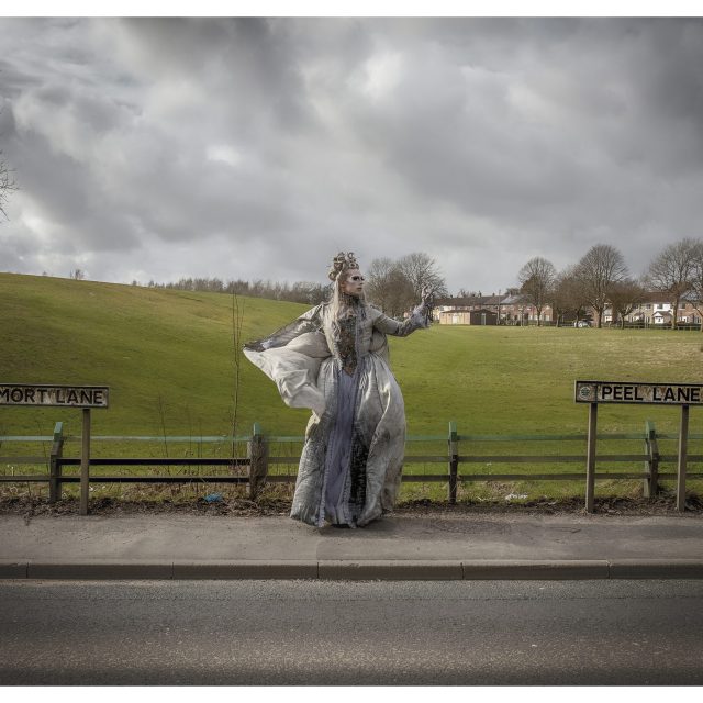 Cheddar Gorgeous stands on the pavement of peel lane with a field behind her, dressed in a grey gown and veil